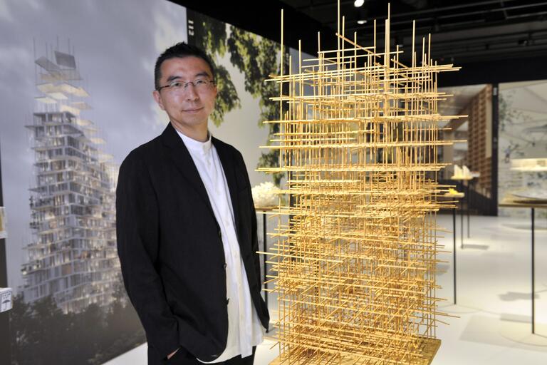 Man in black blazer and white shirt standing next to an intricate geometric architectural model in a gallery.
