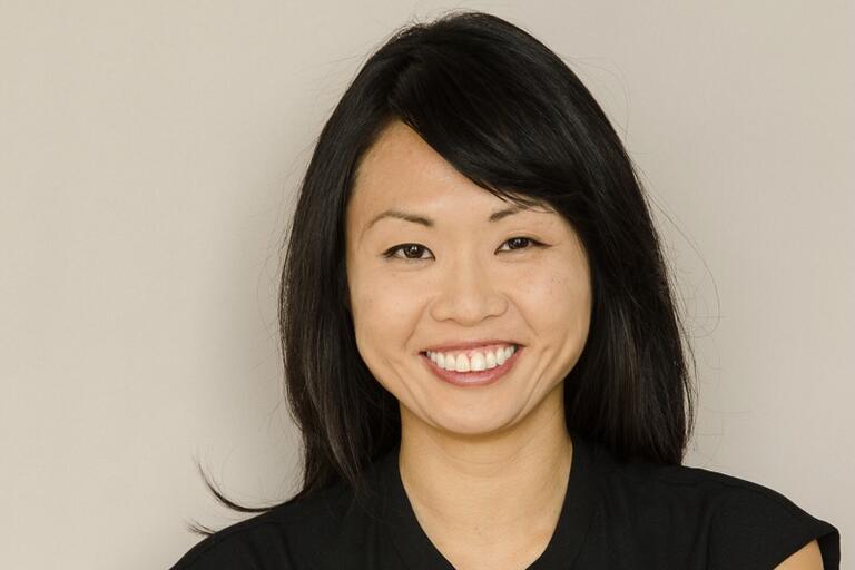 A close-up of a smiling woman with black hair against a light gray background.