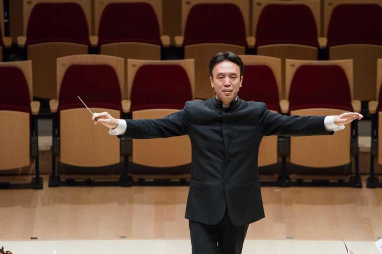 Conductor standing on stage with arms extended, holding a baton. Rows of empty auditorium seats in the background.
