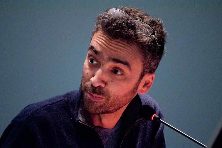 Man with curly hair and a beard speaks into a microphone, wearing glasses on his head and a dark blue sweater.