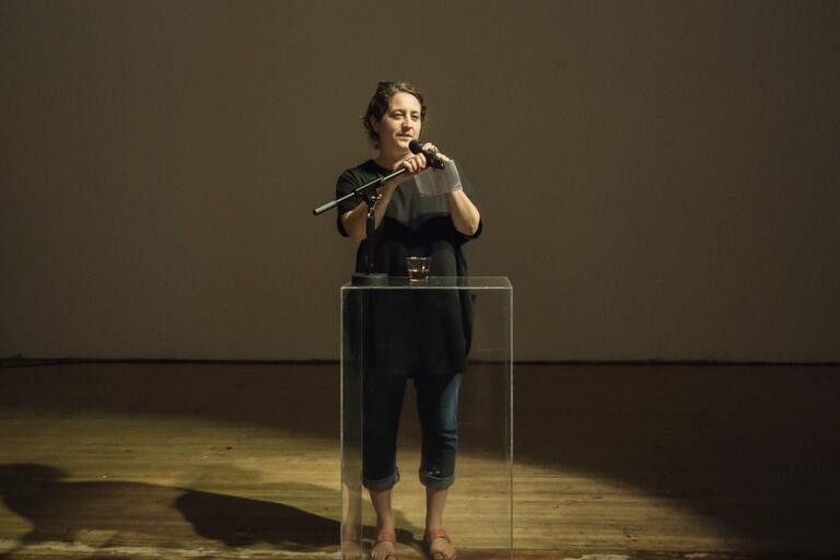 Person speaking at a transparent podium with a microphone in a dimly lit room.