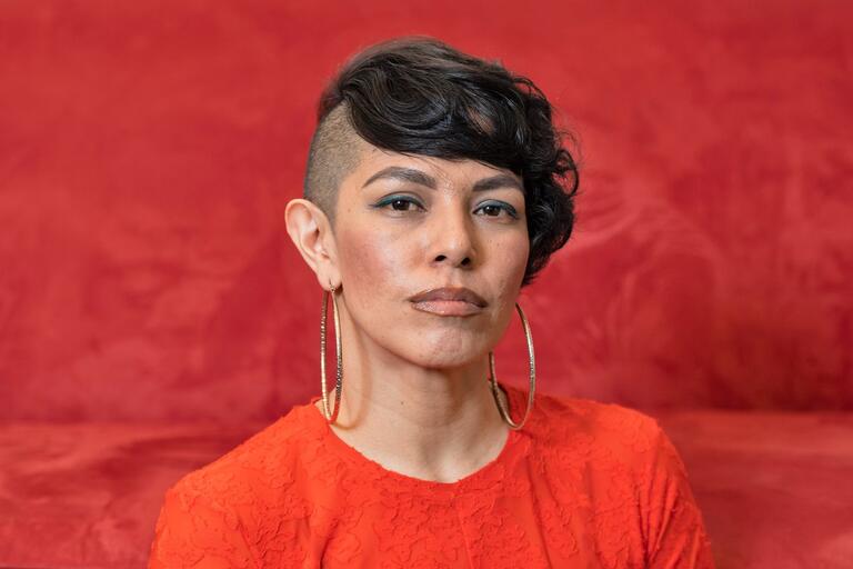 Person with shaved head on one side and short wavy black hair on the other, wearing large gold hoop earrings and a bright red top, against a solid red background.