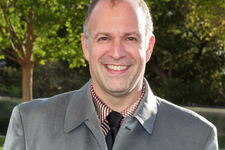 Man smiling in a gray suit jacket, checkered shirt, and black tie, standing outdoors with trees in the background.