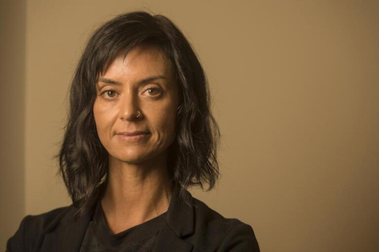 Portrait of a person with medium-length black hair, wearing a black top and jacket, standing against a beige background.