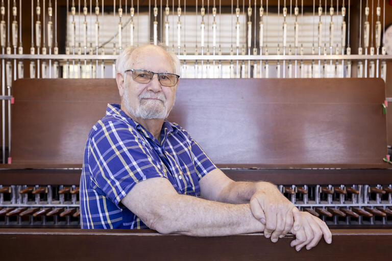 Older man in glasses and blue plaid shirt seated in front of a large wooden and metal musical instrument.