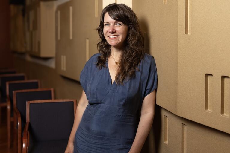 Woman with wavy brown hair in a navy-blue dress stands in front of a wall with beige acoustic panels and black chairs with wooden frames.