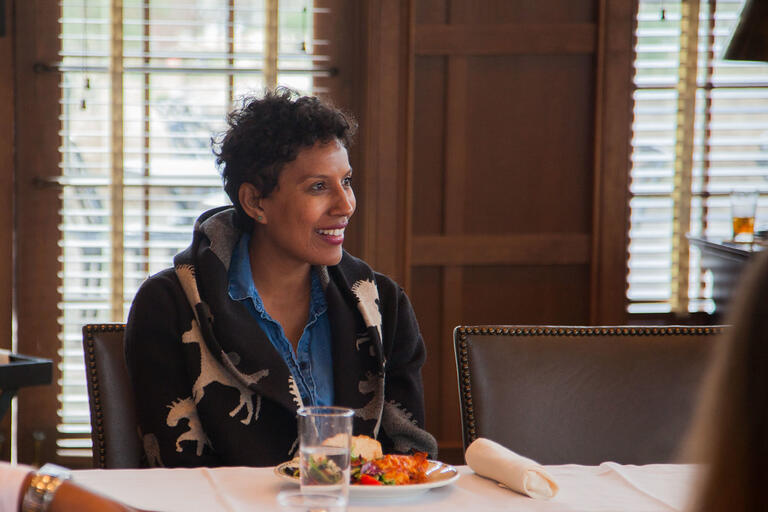 Person smiling while seated at a table set with a glass of water, a plate of food, and a rolled napkin, with wooden window blinds in the background.