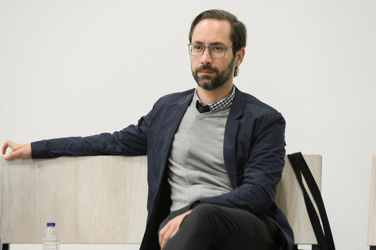 Man seated on a bench with a water bottle beside him, wearing glasses, a dark blue jacket, and a gray sweater over a checkered shirt.