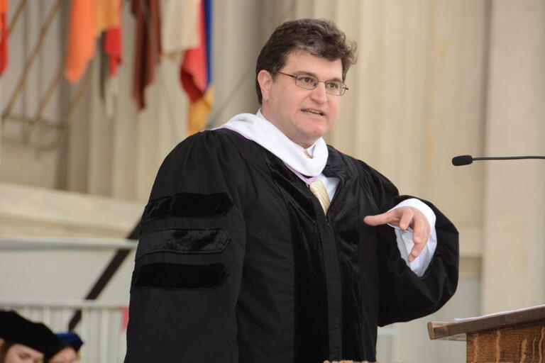 Man in academic regalia speaking at a podium with flags in the background.