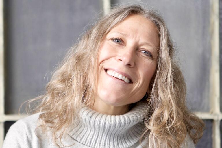 Close-up of a smiling person with wavy blonde hair wearing a gray turtleneck sweater.
