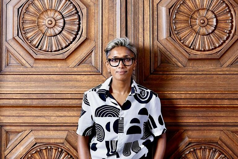 Person standing in front of a carved wooden door with geometric patterns, wearing a patterned short-sleeve shirt and glasses.