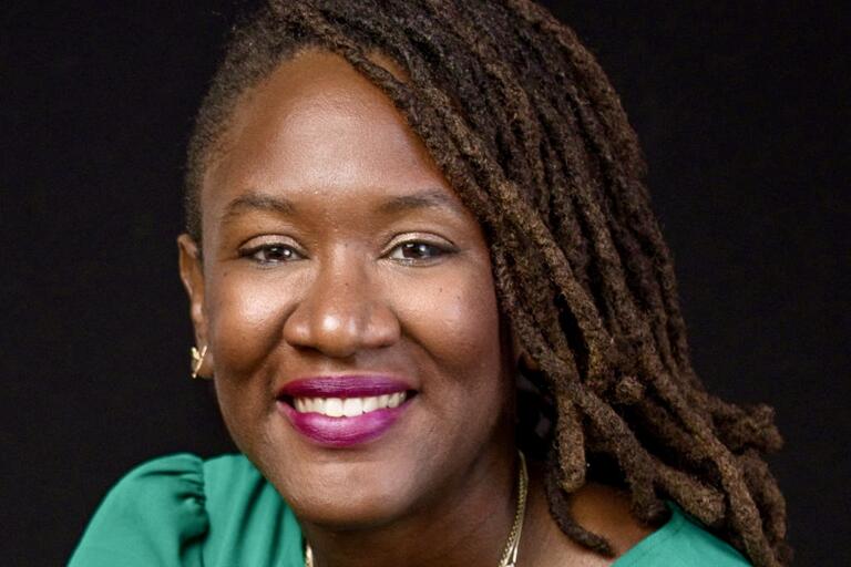 A smiling person with dreadlocks, wearing a green top and gold necklaces, against a black background.