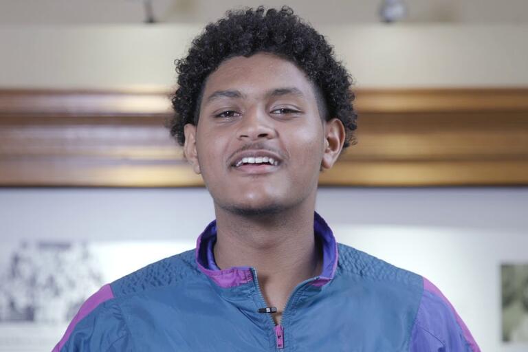 Young person with curly hair in a blue and purple jacket standing indoors with a wooden frame and wall art in the background.