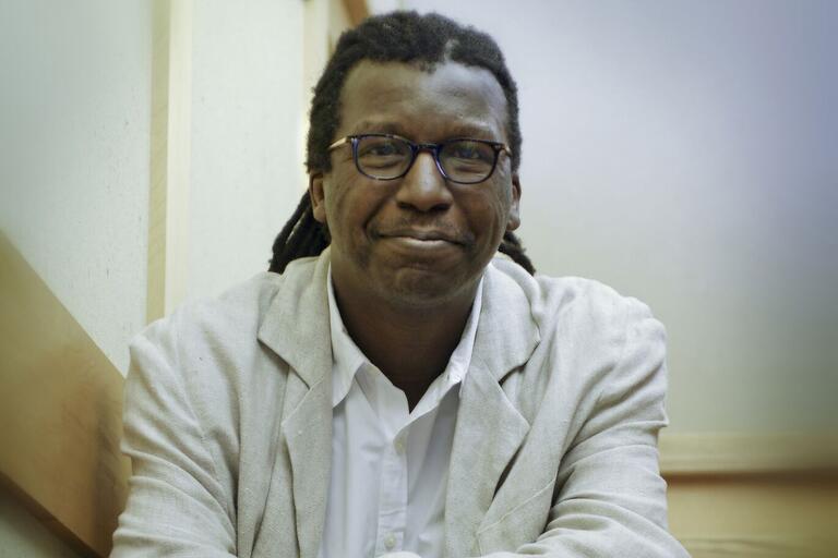 Portrait of a man with glasses, dreadlocks, a light blazer, and a white shirt, looking at the camera.