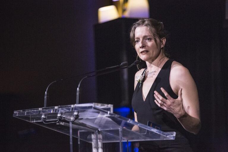 A woman speaking at a podium with microphones, wearing a black sleeveless dress.