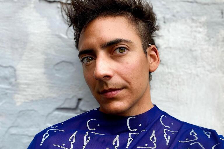 Close-up portrait of a person with short, spiky dark hair, wearing a blue shirt with abstract white patterns against a gray textured wall.