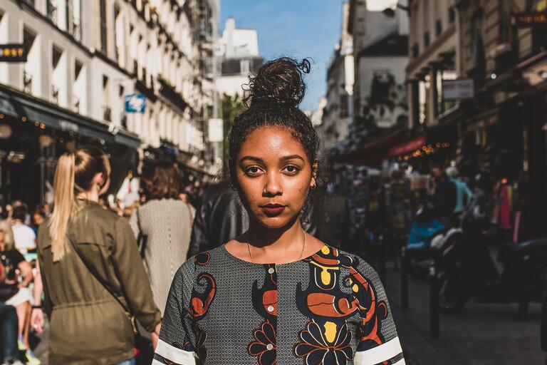 Woman with hair in a bun, wearing a patterned dress, stands on a busy urban street.