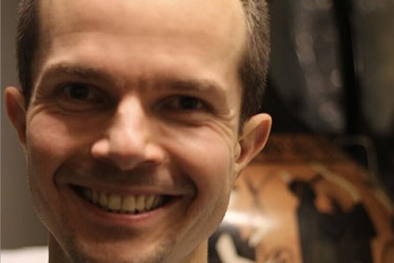 Close-up of a person smiling with a blurry background featuring a pattern resembling an ancient Greek vase design.