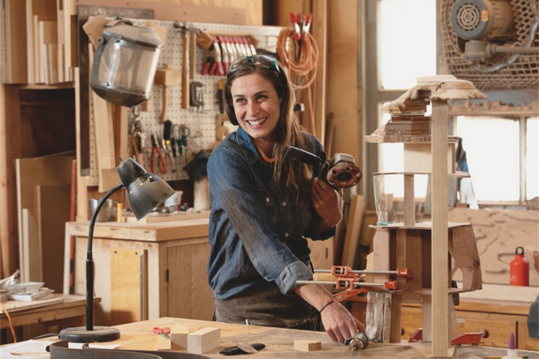 Woman in a woodworking shop holding a power drill and smiling.