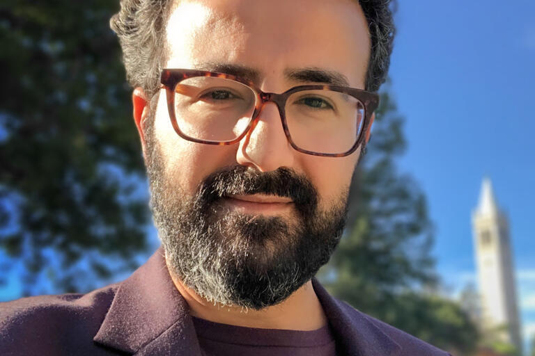 A man with dark hair and beard wears glasses & a purple suit, looks into camera