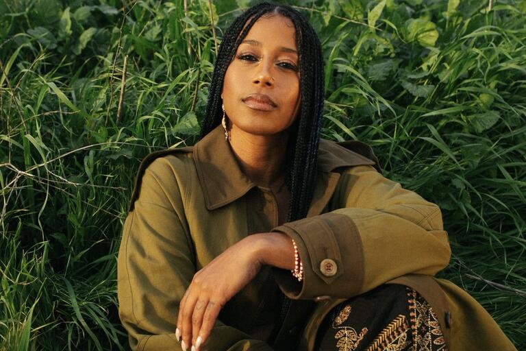 Person with long braids sitting in dense green foliage, wearing an olive-green coat and a beaded bracelet.
