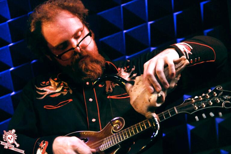 Bearded man holding a skull with antlers and playing a mandolin in front of a textured blue and black wall.
