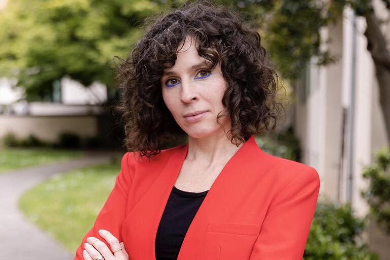 Person with curly hair in a red blazer and black top standing outdoors with arms crossed.