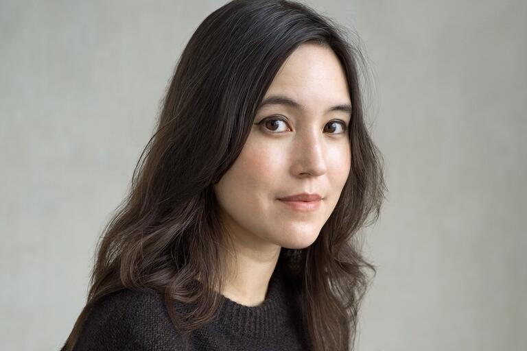 Close-up portrait of a person with dark hair and brown eyes, wearing a dark sweater against a light background.