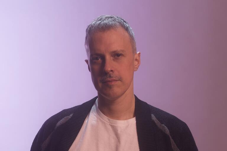 Portrait of a person with short hair wearing a white t-shirt and dark cardigan against a soft purple background.