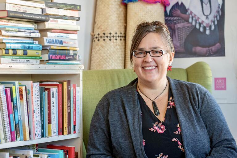 A smiling woman with glasses, seated next to a bookshelf filled with books, with woven mats and artwork in the background.