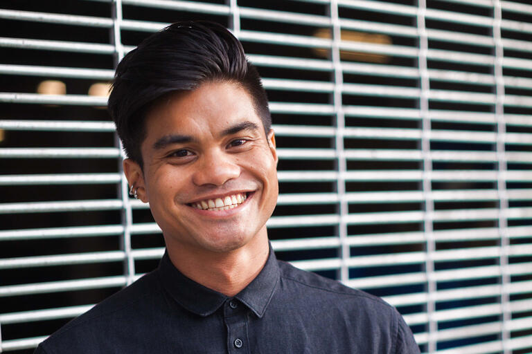 Person smiling in front of a wall of horizontal metal bars, wearing a dark button-up shirt.
