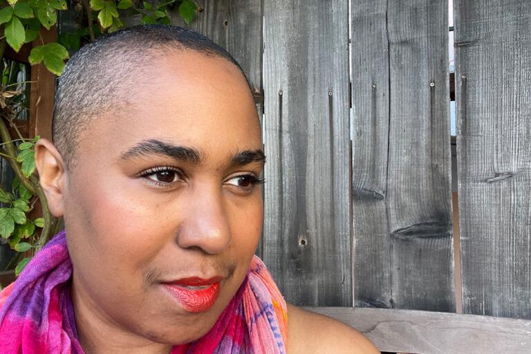Close-up of a person with a shaved head, bright red lipstick, and a colorful scarf, standing in front of a wooden fence.