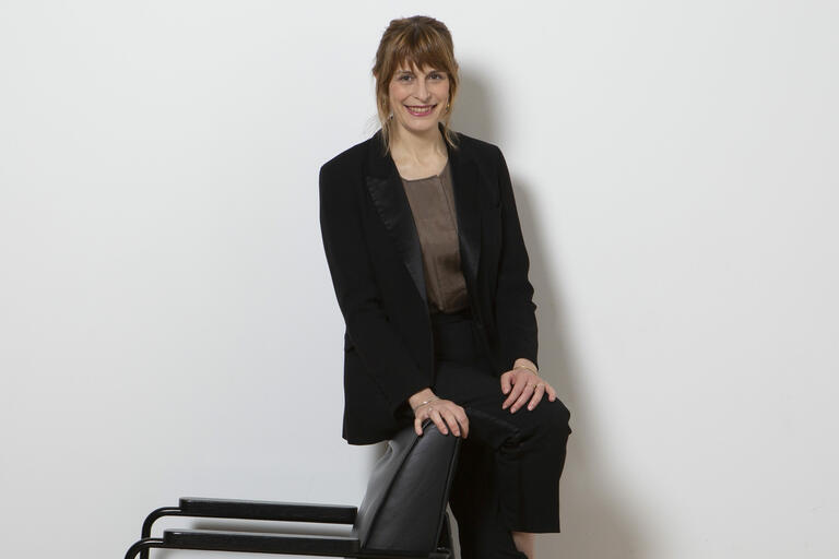 Woman in a black suit leaning against a black chair with a white background.