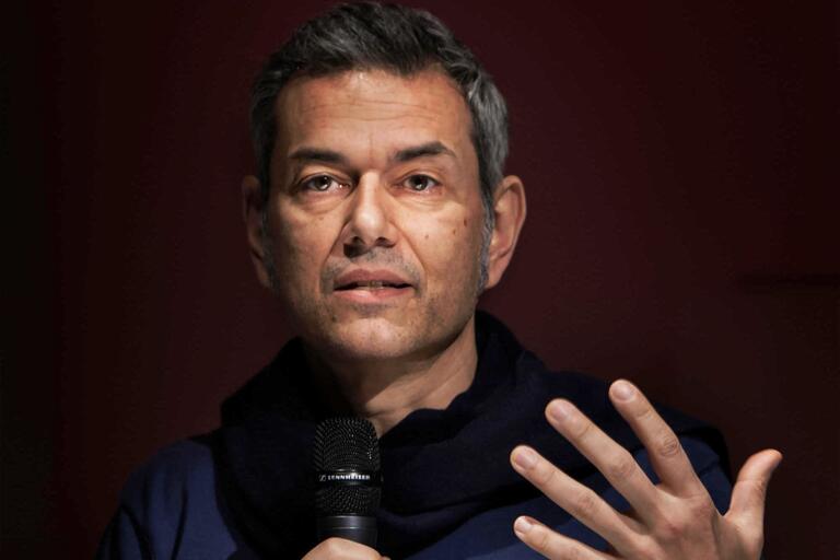 Close-up of a middle-aged person speaking into a microphone with a deep red background.