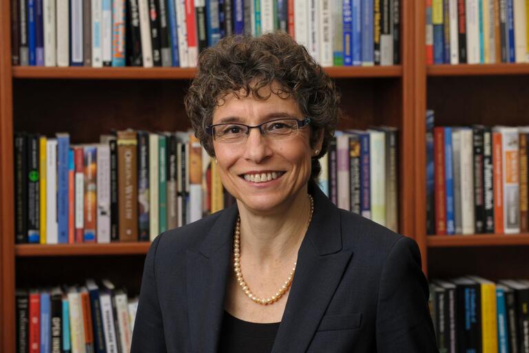 Person in a dark suit with pearls standing in front of a bookshelf filled with books.