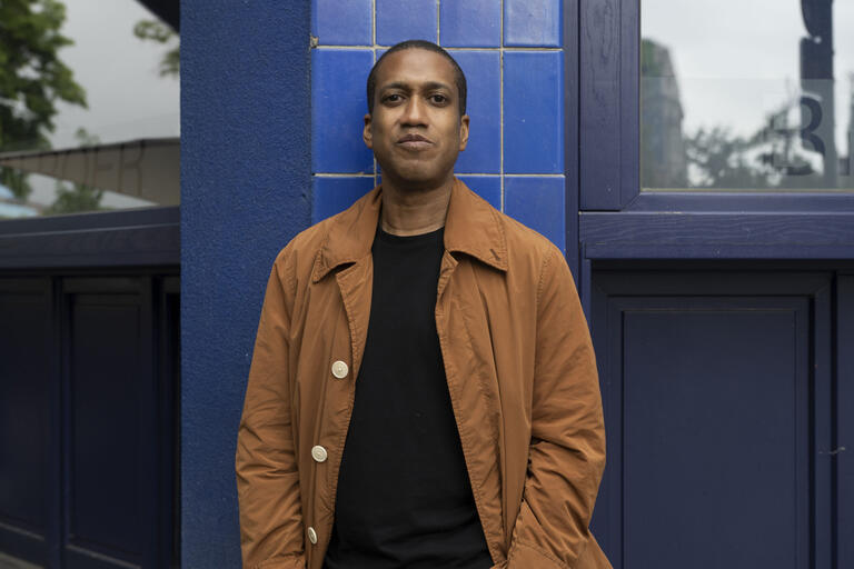 Person wearing a tan trench coat and black shirt stands in front of a blue-tiled building.
