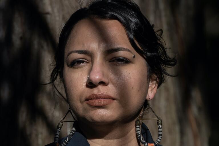 A person wearing a navy blue shirt and large striped hoop earrings stands in front of a tree trunk with shadows cast over their face.