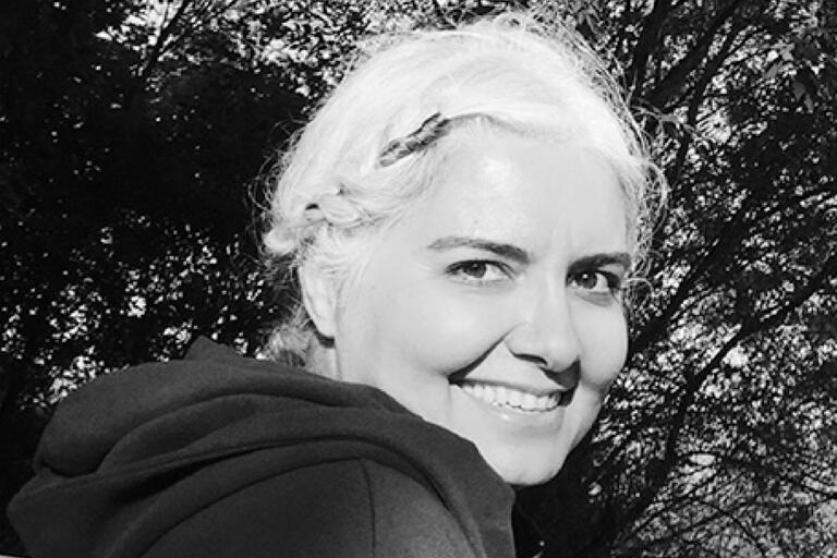Black and white photo of a smiling person with light-colored hair in an outdoor setting with tree foliage in the background.