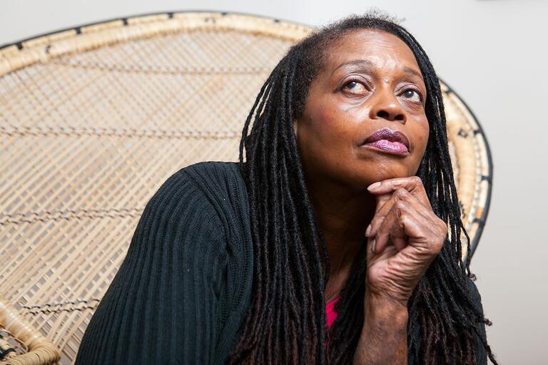 Thoughtful older woman with long dreadlocks seated in front of a woven wicker chair, resting her chin on her hand.