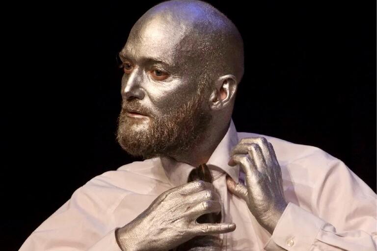 Person with silver-painted face and hands adjusting a tie against a black background.