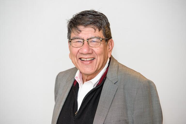 Older man with glasses smiling, wearing a gray blazer and black zippered sweater over a white collared shirt.