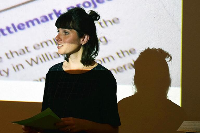 A woman speaking in front of a projected document on a wall.