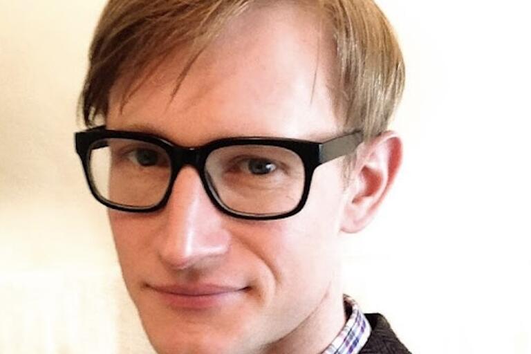 Close-up portrait of a young man with short light brown hair, wearing black-rimmed glasses and a checkered shirt.