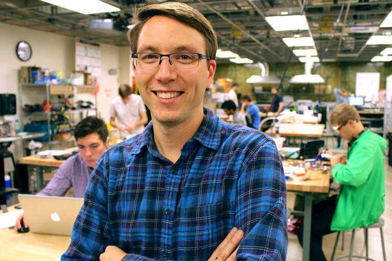 A man with glasses and a flannel shirt stands smiling in a busy, well-lit workshop with several people working in the background.