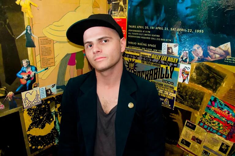 Man in black cap and dark blazer with a pin stands in front of a wall covered with various posters and flyers.