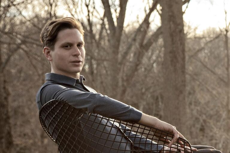 Person seated on a wire mesh chair in a woodland setting with leafless trees.