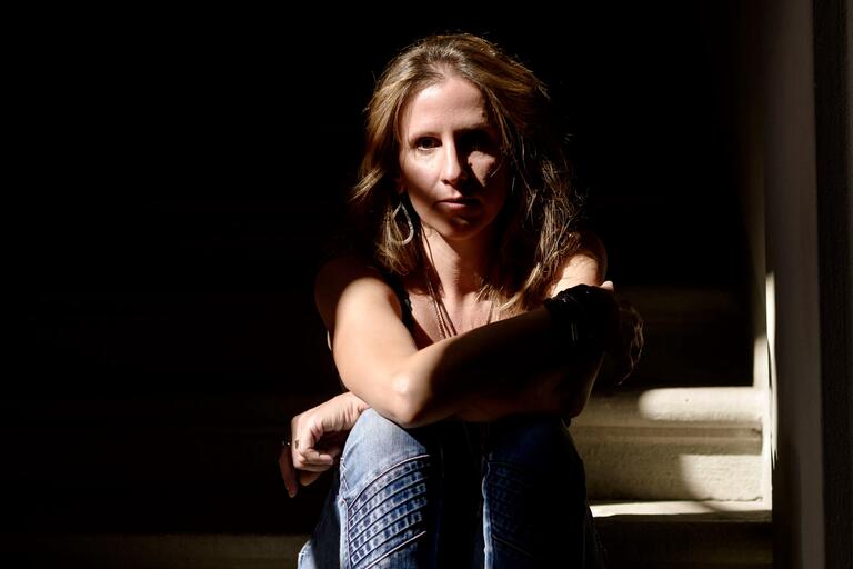 Woman with wavy hair sits on a stairway in partial sunlight, arms resting on her knees.