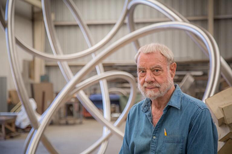 Older man in a denim shirt in front of large metallic structure in a workshop.