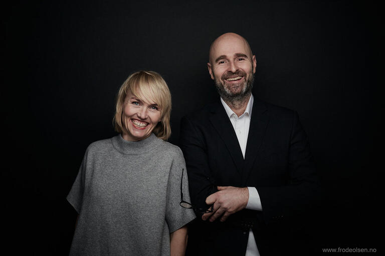 Two smiling people standing against a black background, the woman in a gray top and the man in a dark suit with a white shirt.