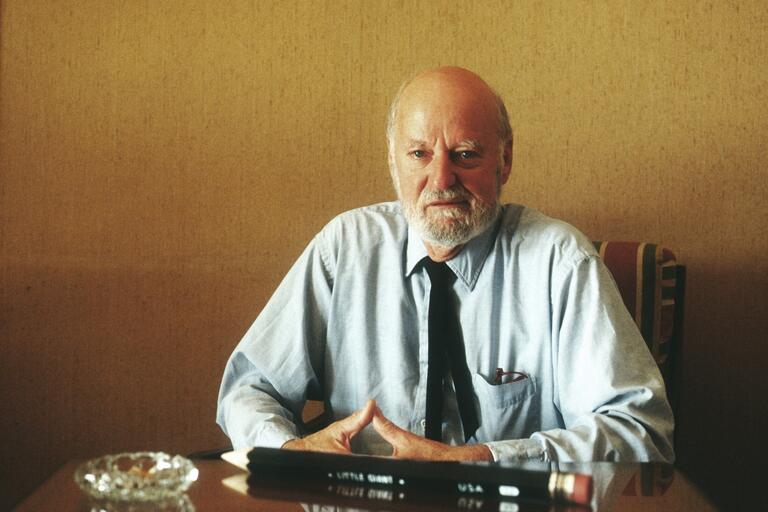 Older man with a white beard wearing a blue shirt and black tie, sitting at a table with large pencils and an ashtray.